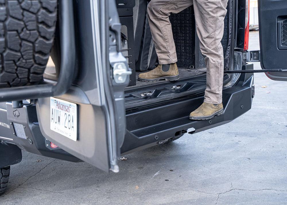 Side profile of a Mercedes Sprinter van with the rear cargo doors open, showing a person standing on the integrated step of the ROAMBUILT Rear Bumper. The image highlights the bumper’s sturdy aluminum construction, secure footing, and practical design for easy access to the cargo area.
