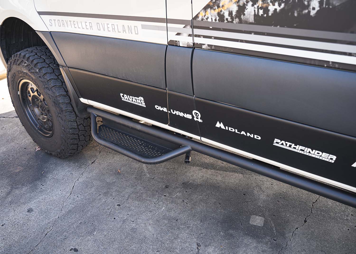 Driver side view looking from the rear toward the front, showcasing the full ROAMBUILT Mercedes Sprinter Side Step mounted under the van with visible perforated aluminum decking 