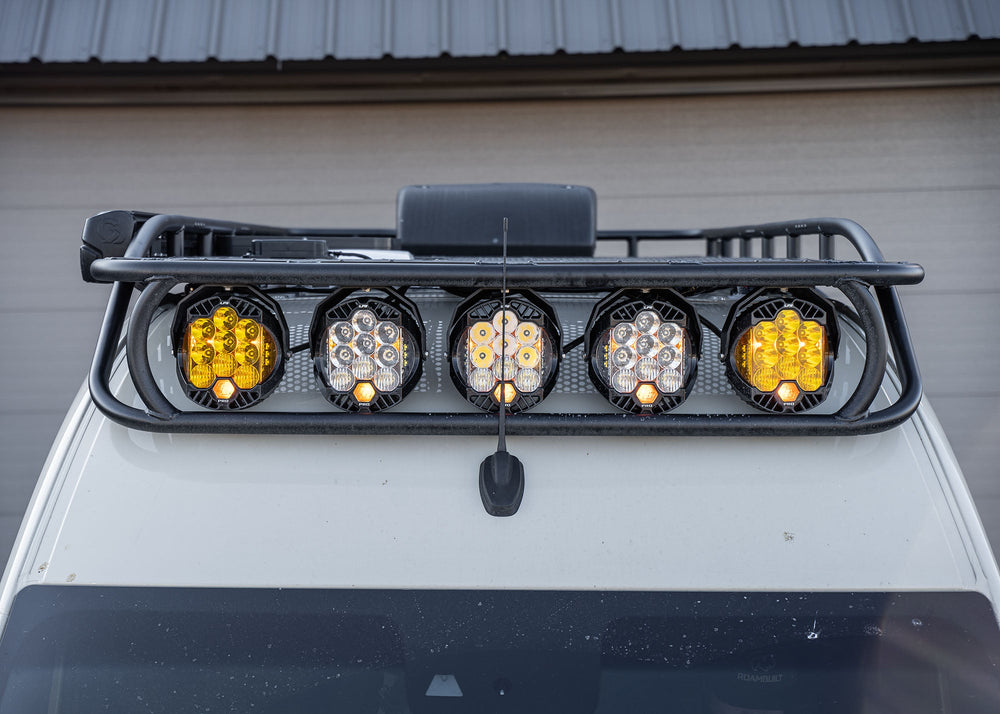 Close-up front view of the ROAMBUILT Safari 2.0 roof rack showing Baja Designs LP9 lights securely mounted, highlighting the curved front design on a 2007+ Mercedes Sprinter.