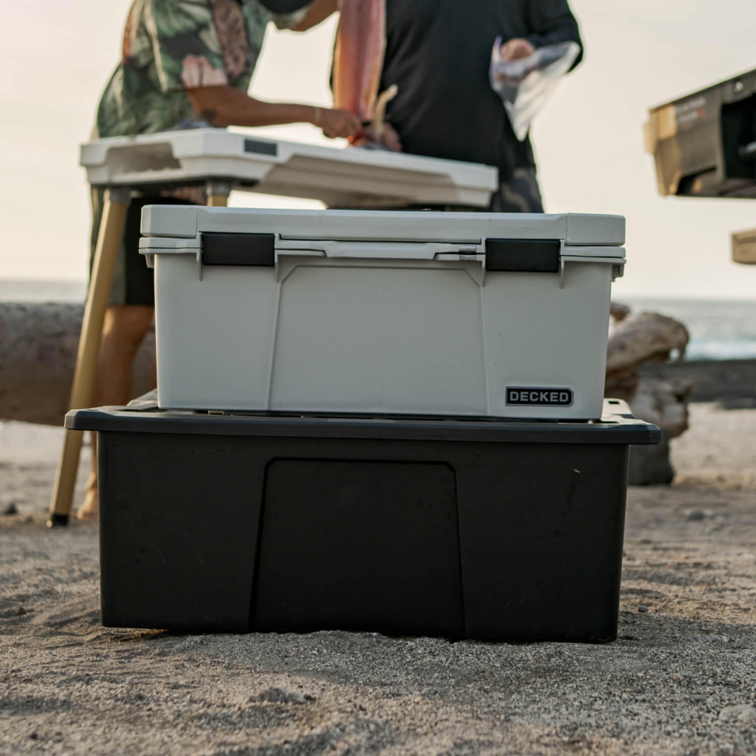 [A0010-SIXR-GRY] The Cool Gray Sixer stacked on top of a Payloader 32 D-co Case, in the background are some friends filleting a fish for a quick dinner on the beach.