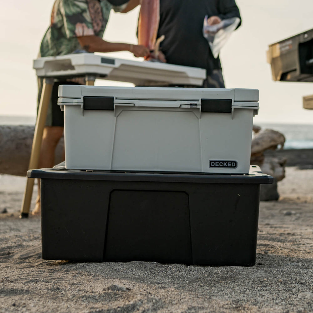 [A0010-SIXR-GRY] The Cool Gray Sixer stacked on top of a Payloader 32 D-co Case, in the background are some friends filleting a fish for a quick dinner on the beach.