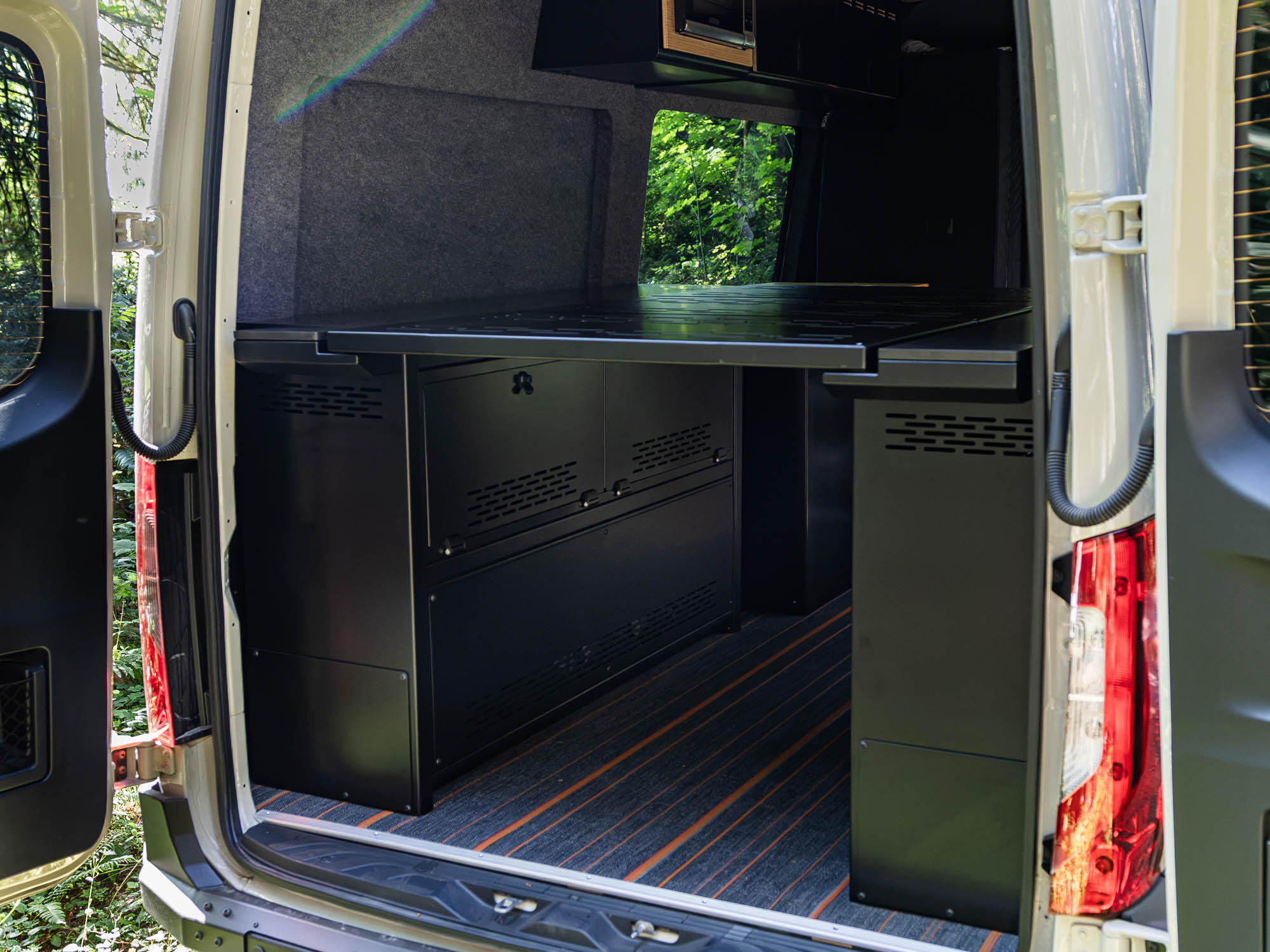 Angled view of installed Modular Cabinet Bed System for Sprinter & Transit in North-South orientation from rear of van, showing cabinet doors and fixed panel in use