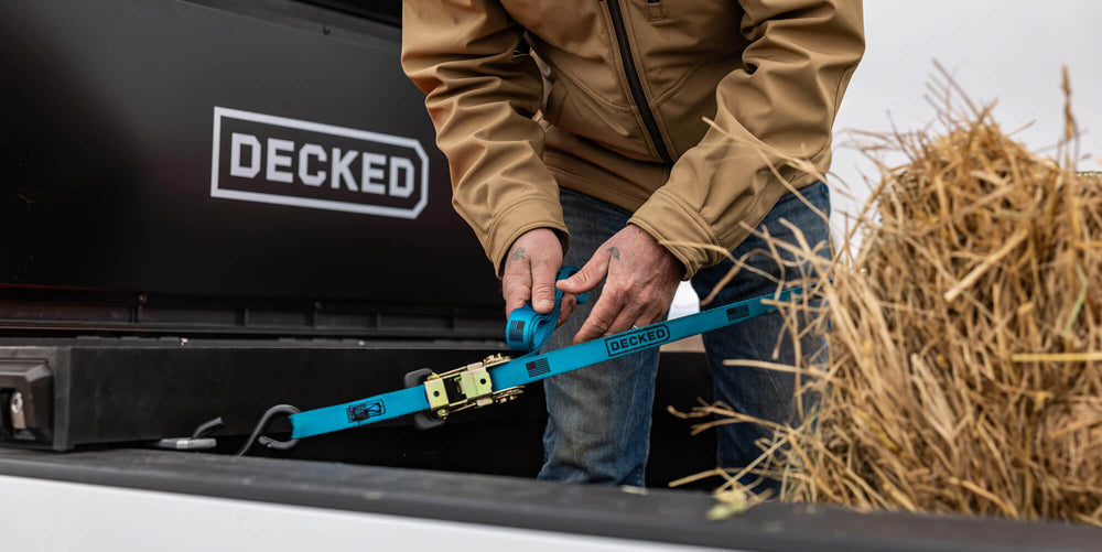 @expanded [ATB-DRING] Person securing a bale of hay with a blue strap next to a DECKED'Tool Box.