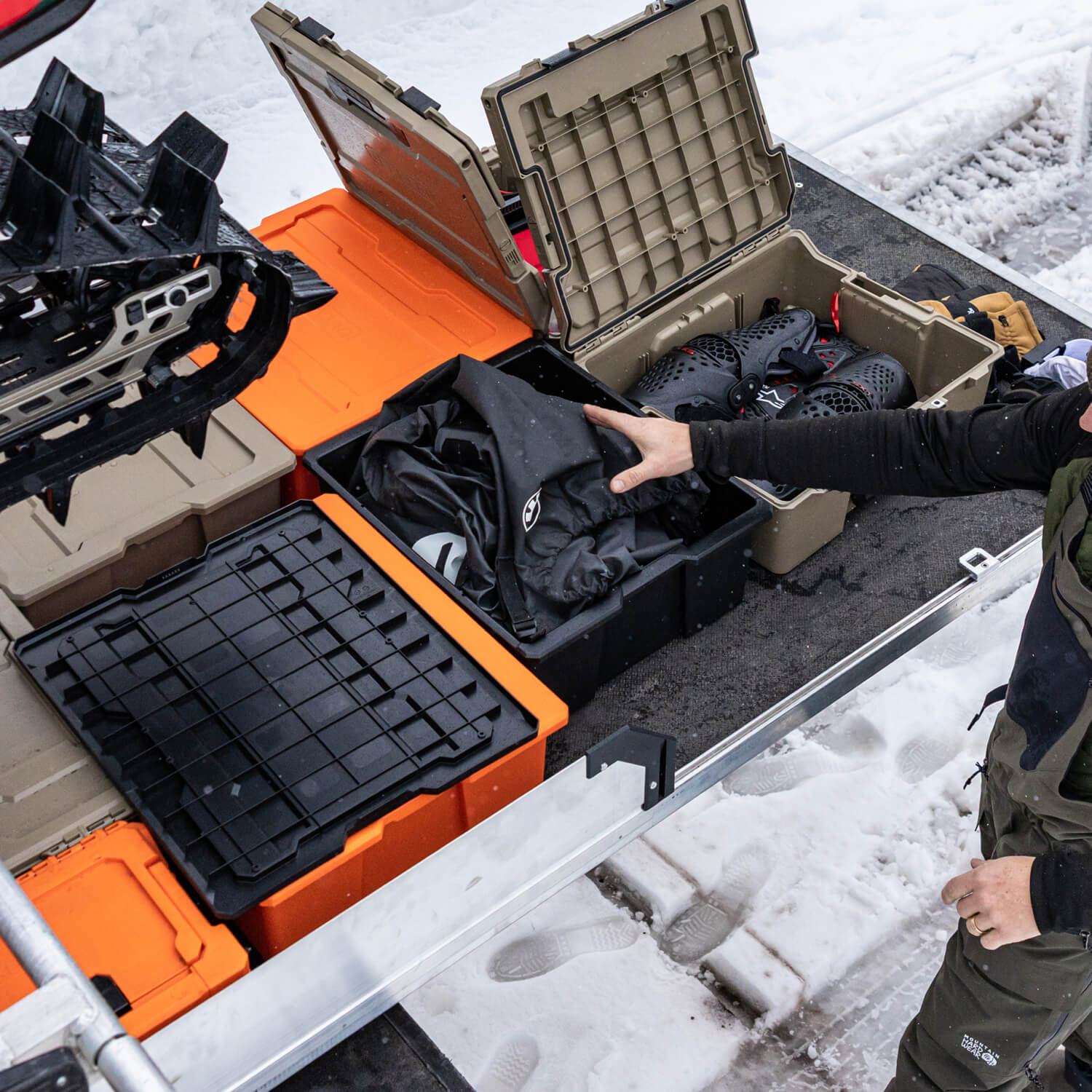 [A0041-DBIN-BLK, A0040-DBIN-BLK] Person organizing snowmobiling gear in a Payloader case on top of a DECKED CargoGlide with snow-covered ground in the background