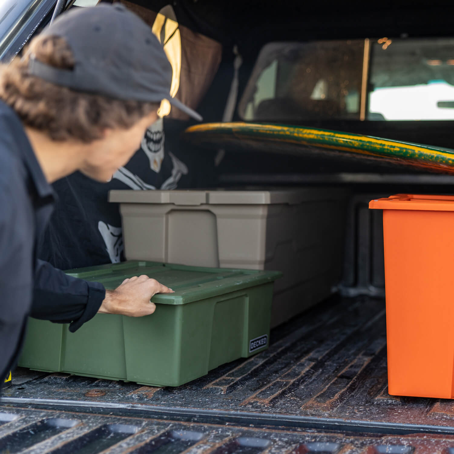 [A0041-DBIN-GRN, A0040-DBIN-GRN] A man loading Payloader Bins into the bed of his truck along with a surfboard for a day on the water.