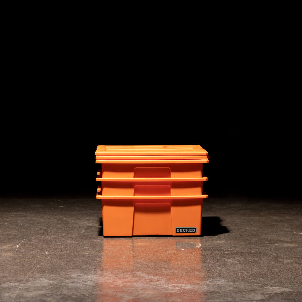 [A0041-DBIN-ORG, A0040-DBIN-ORG] Three blaze orange Payloader Bins nested together with the lids sitting on top in a garage with a dark, shadowy background.
