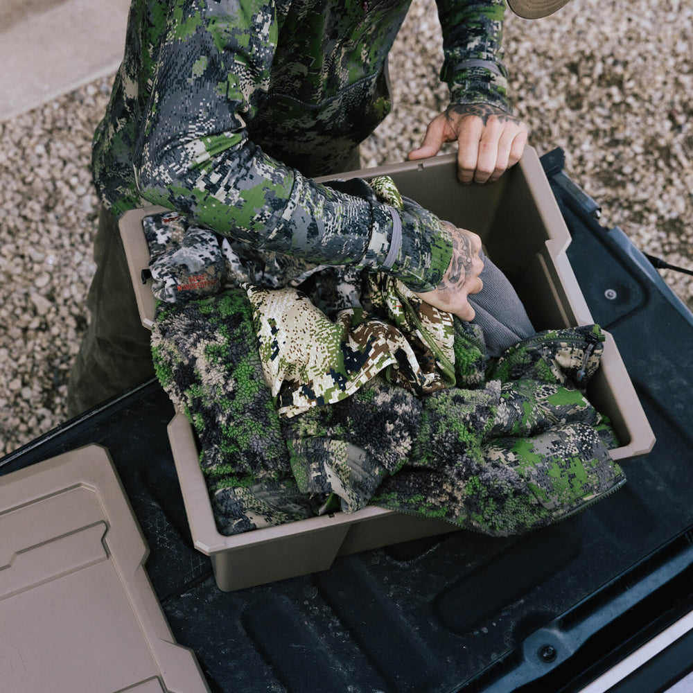 [A0041-DBIN-TAN, A0040-DBIN-TAN] A man dressed in camo, packing his hunting outerwear into a desert tan Payloader Bin.