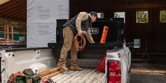 @expanded [DTB2-BLK, DTB2T22-BLK] Person loading a black DECKED Tool Box in their truck bed with supplies, including a hose and a Halfrack D-co Case.