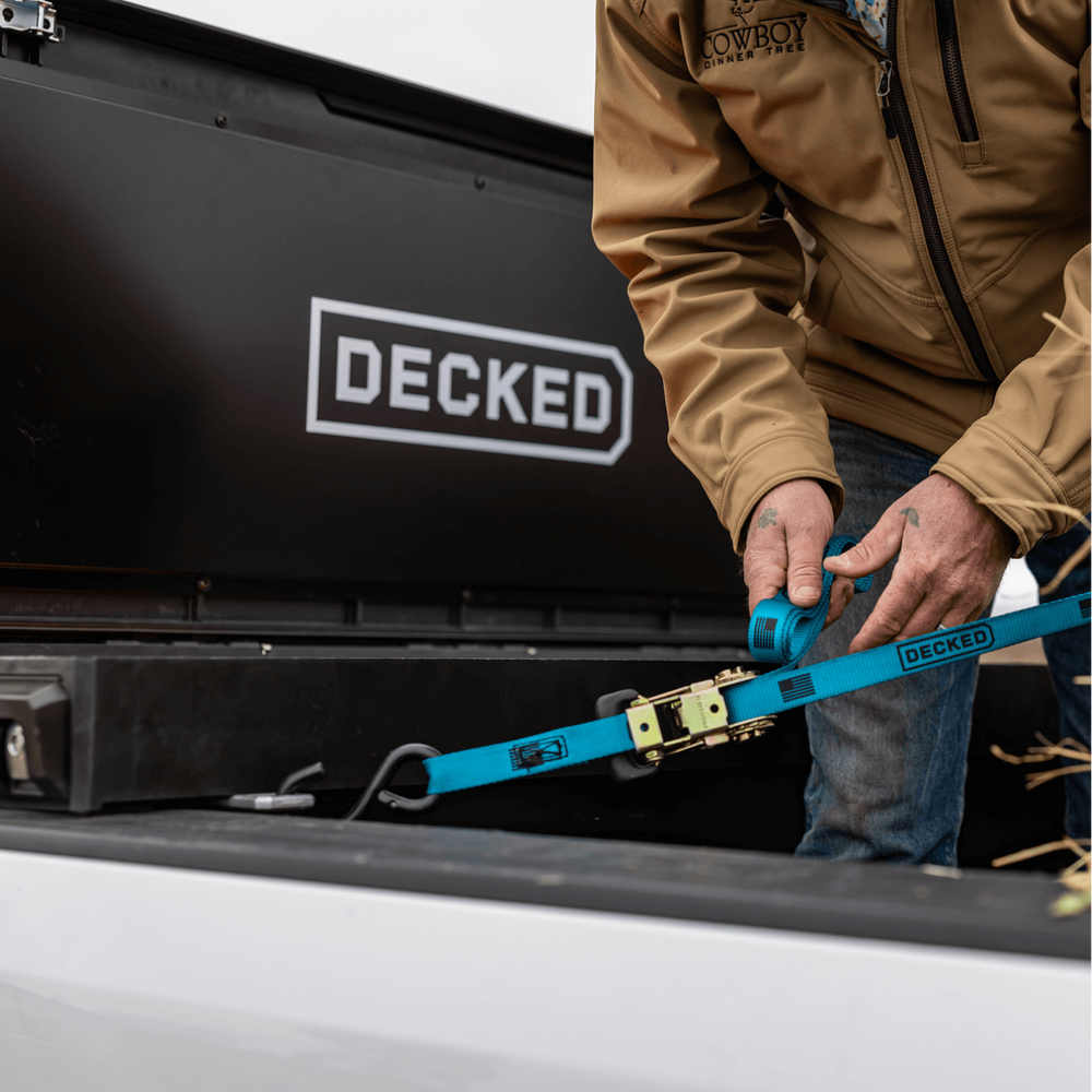 [DTB2-BLK, DTB2T22-BLK] Person securing a DECKED blue Ratchet Strap to their DECKED Tool Box in order to lash down the cargo in their truck bed.
