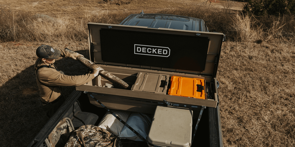 @expanded [DTB2-TAN, DTB2T22-TAN] Person loading a shotgun case into their 'DECKED' Tool Box storage system in an outdoor setting.